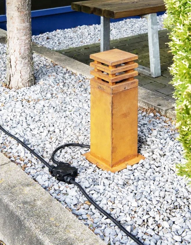 A black outdoor electrical junction box with a cable running into it, connected to a garden light fixture on a bed of