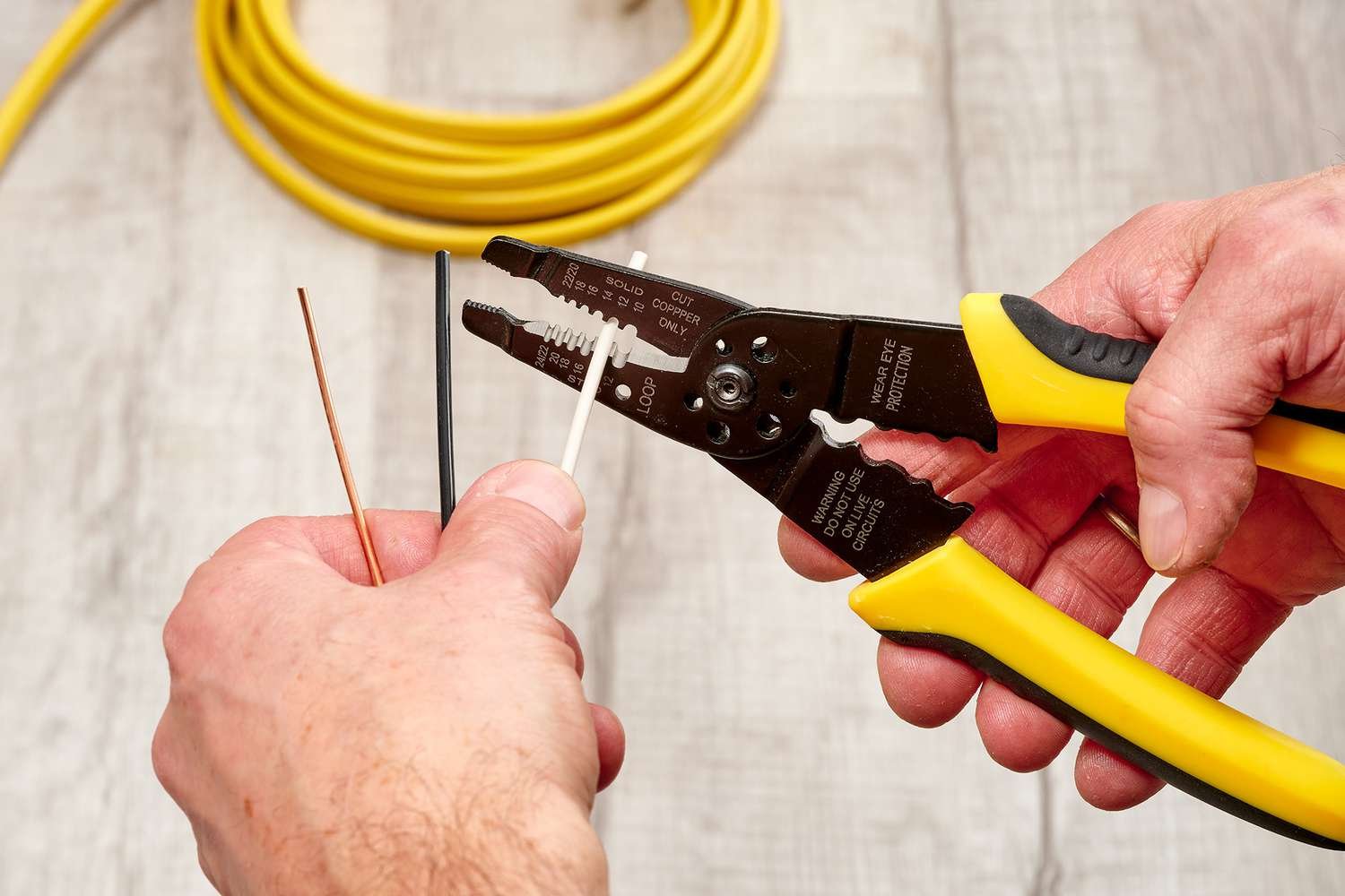 A pair of hands using a manual Electrical Wire Stripper on a black and a white insulated wire