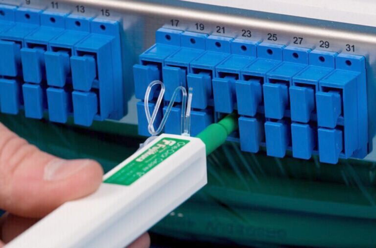 A hand using an Optical Fiber Cleaner pen to clean an LC fiber optic connector in a blue patch panel.