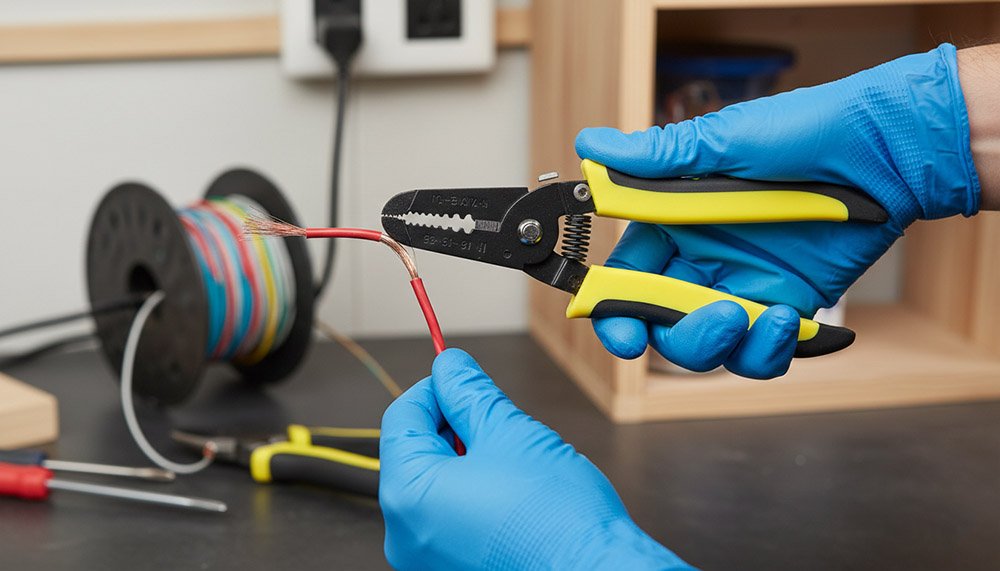 A gloved technician using a wire stripper cutter to remove insulation from a red electrical cable in a workshop setting